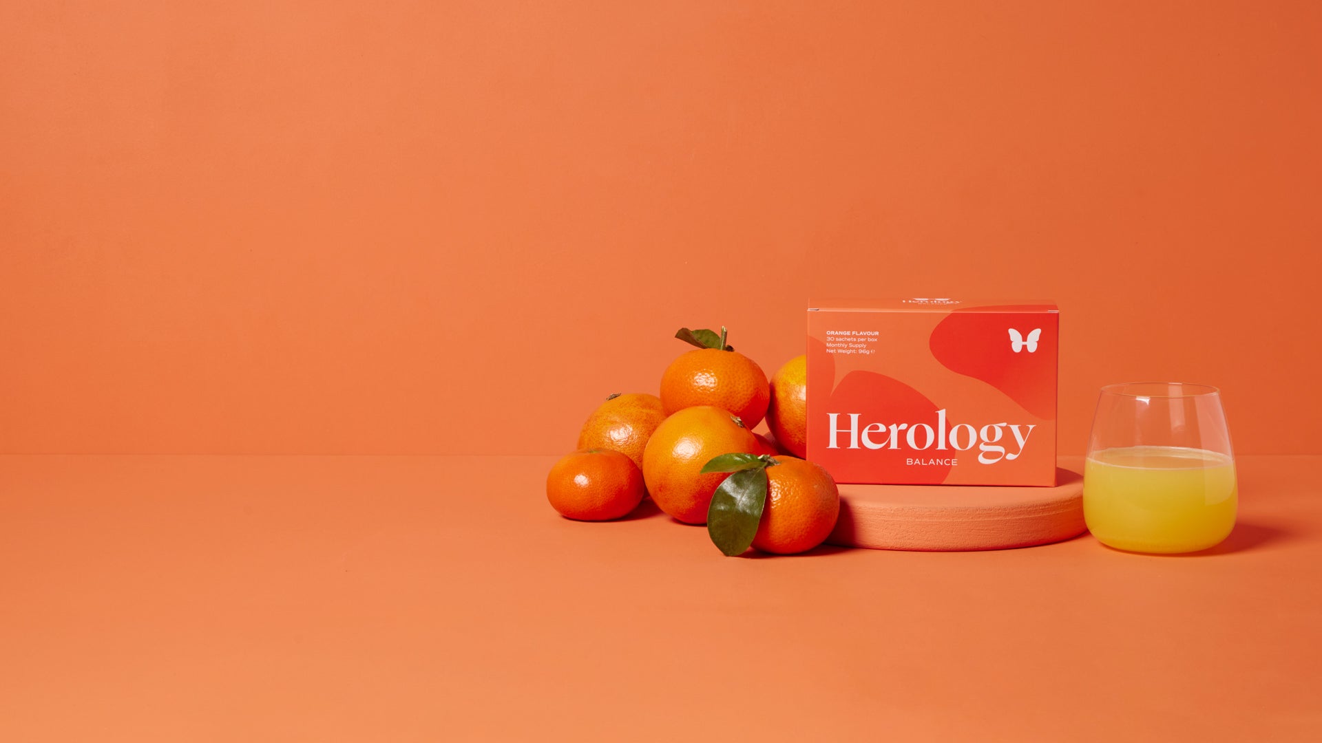 Orange still life featuring a Herology Balance box, oranges, and a glass of juice against an orange backdrop.