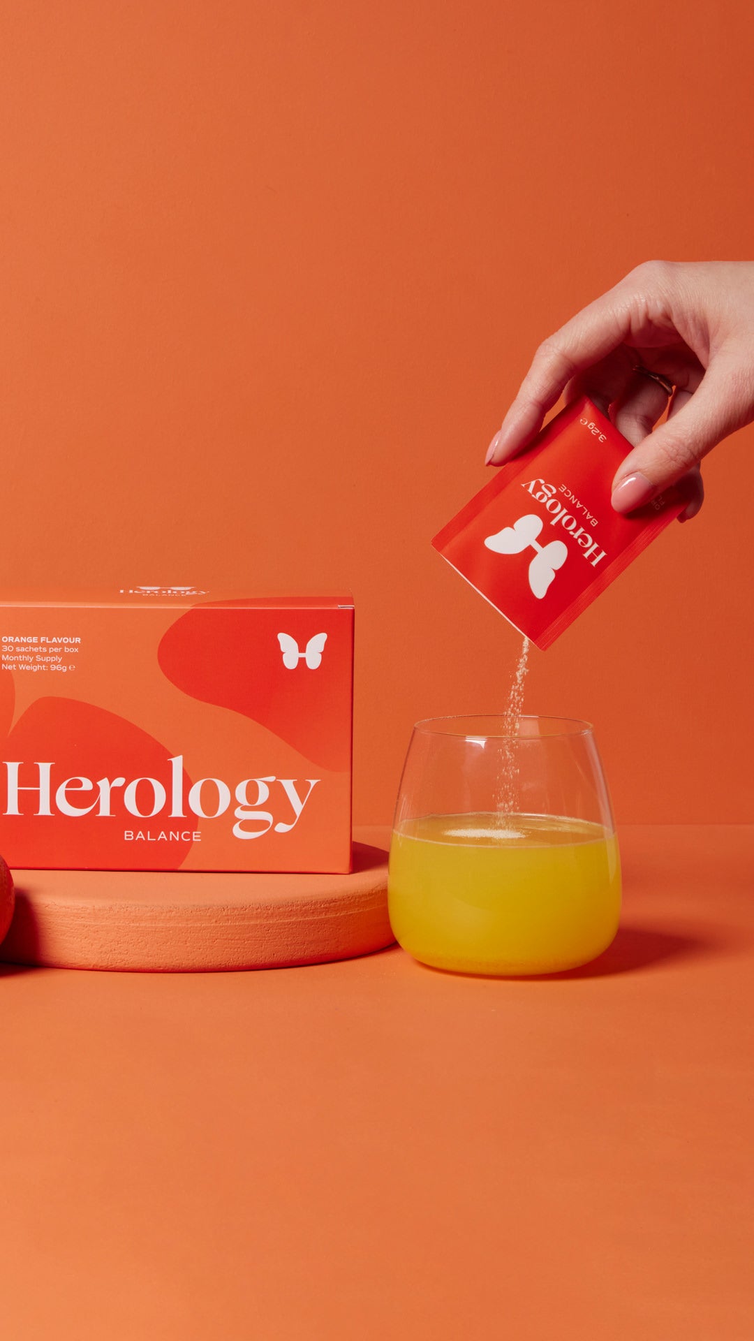 A hand pours the contents of a Herology Balance packet into a glass of orange liquid, next to a box of the product, against an orange background.