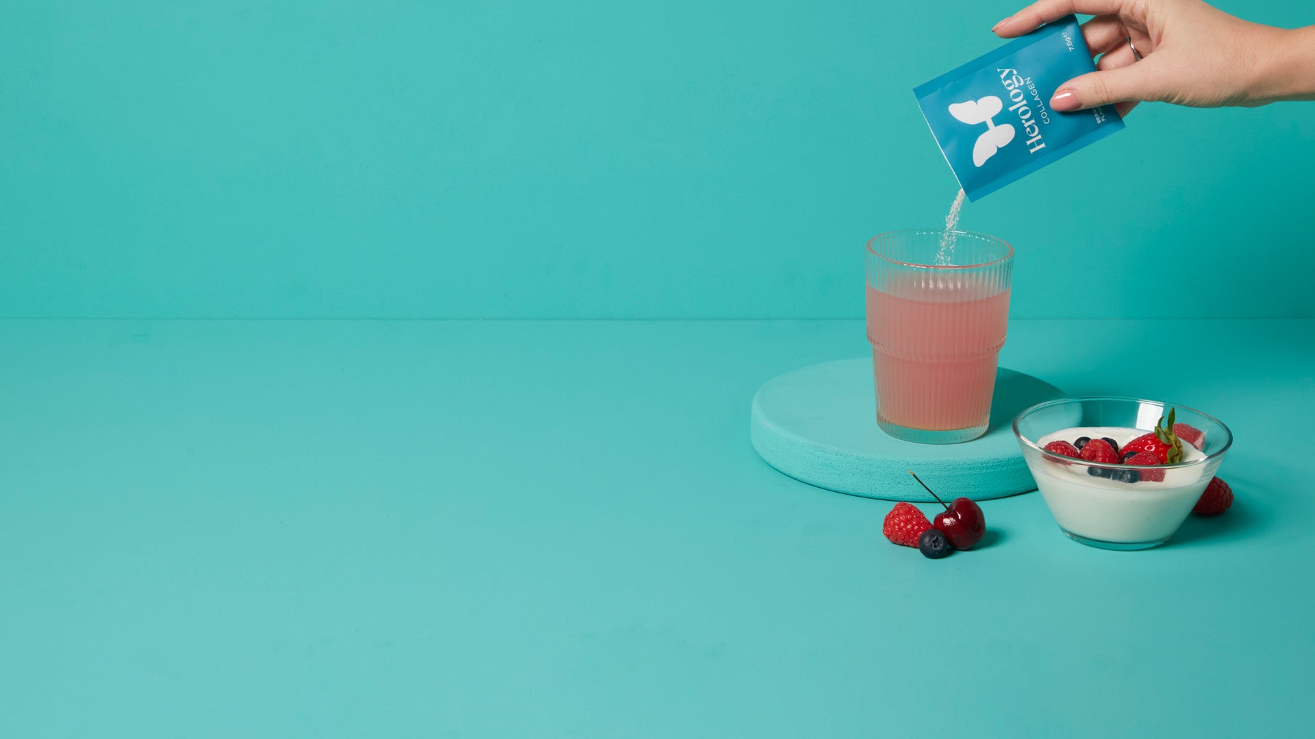 Hand pouring Herology collagen powder into a glass of liquid, with yogurt and berries nearby, on a turquoise background.