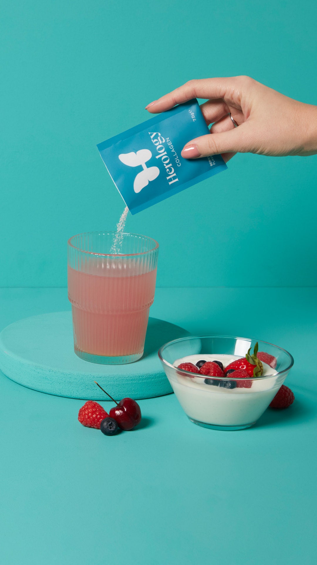A hand pours Herology collagen powder into a glass with a pink drink next to a bowl of yogurt and berries on a blue surface.