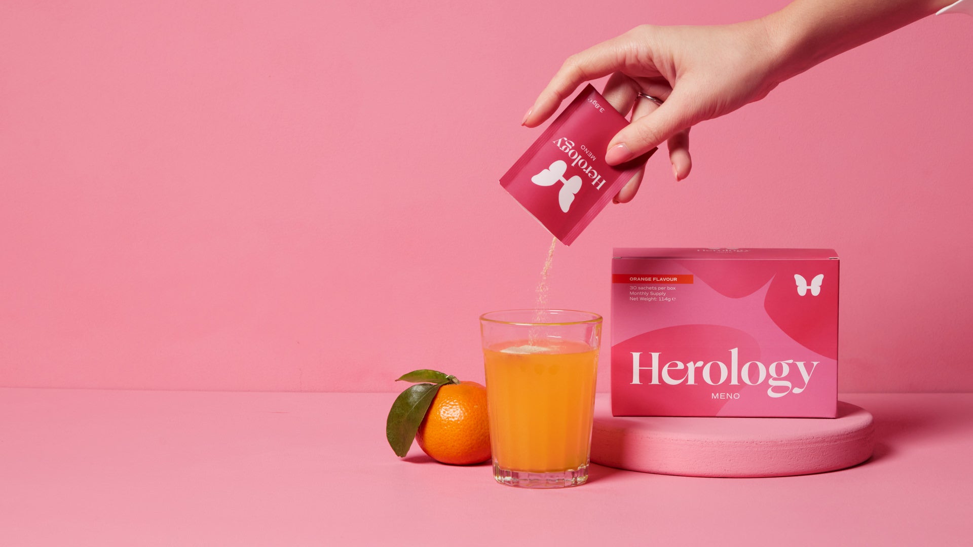 A hand pours contents from a Herology Meno packet into a glass of orange liquid next to the box and an orange.