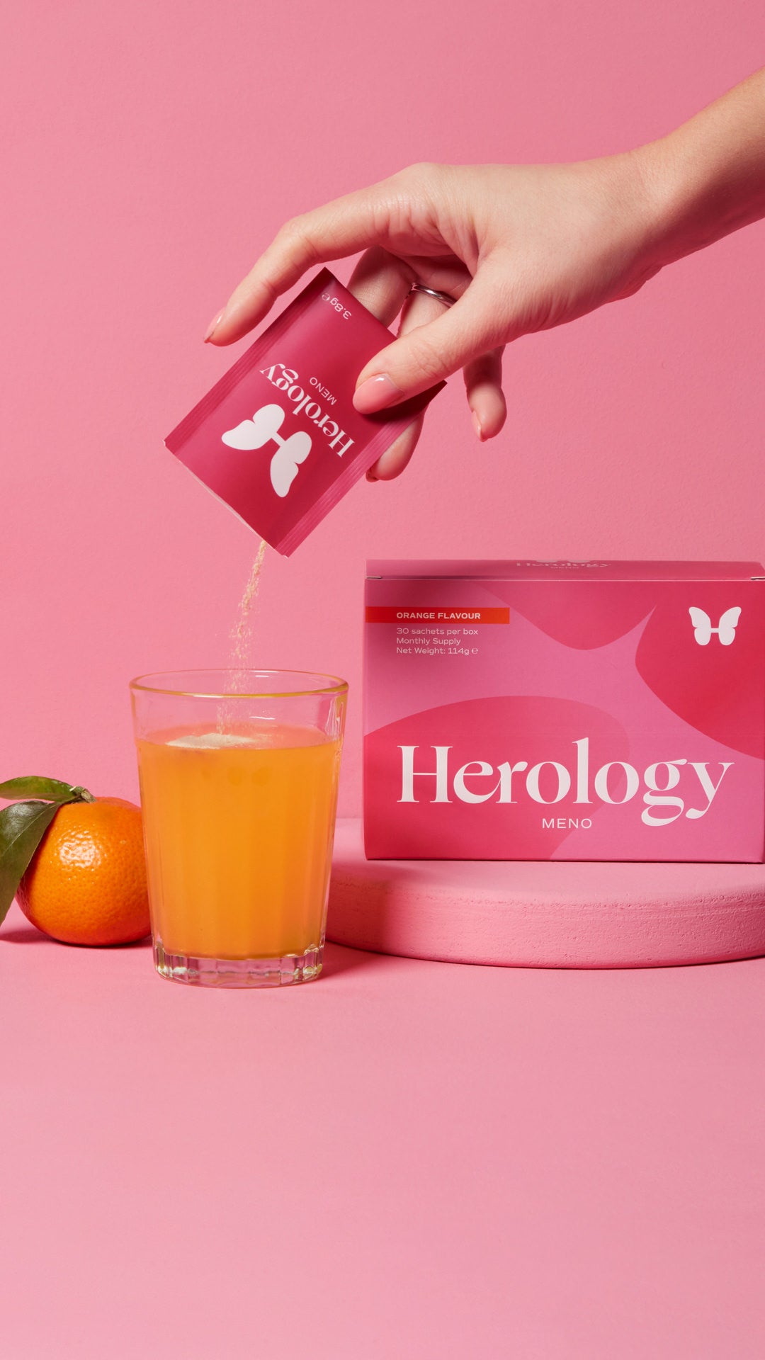 A hand pours contents from a Herology Meno packet into a glass of orange liquid next to the box and an orange.