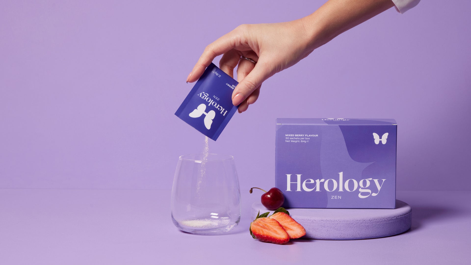 A hand pours Herology Zen mixed berry flavor powder into a glass beside its packaging and a strawberry slice and cherry, all on a purple background.