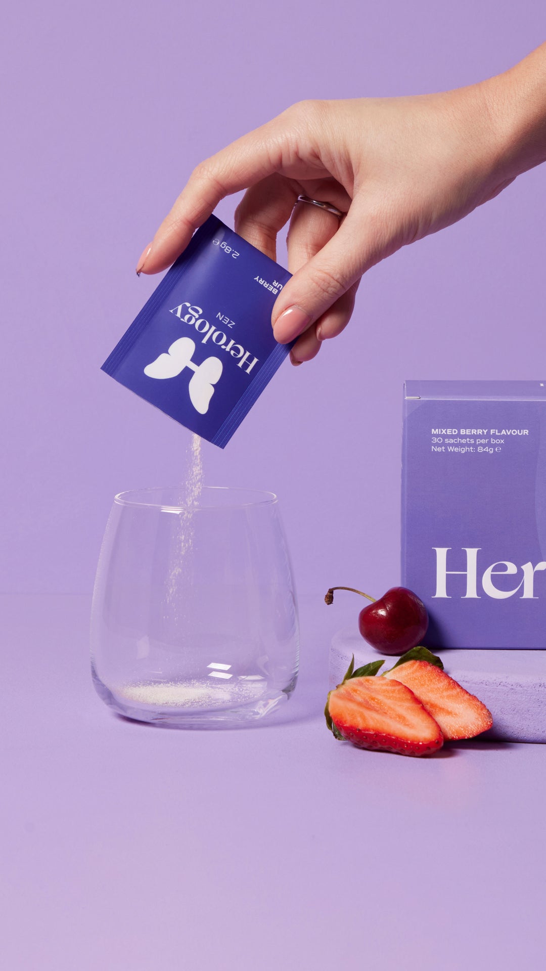 A hand pours Herology Zen mixed berry flavor powder into a glass, with the product box, strawberries, and a cherry nearby, on a purple background.